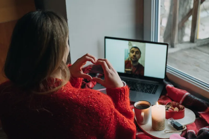 Młoda kobieta w czerwonym swetrze rozmawia przez laptopa z chłopakiem i układa dłonie w kształt serca.